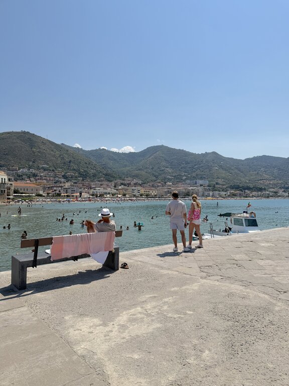 A man and a couple sitting on stone benches overlooking the historic beach and turquoise sea in Cefalù, Sicily