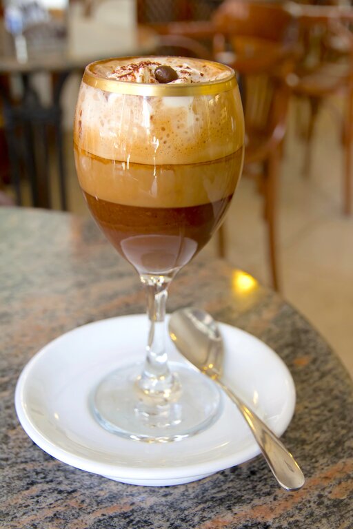 A clear, handleless glass of Bicerin, a traditional Turin coffee drink, showing three distinct layers: thick dark hot chocolate on the bottom, a shot of espresso in the middle and a topping of hand-whisked cream, served on a saucer in a historic Belle Époque cafe ©Morenovel/Depositphotos