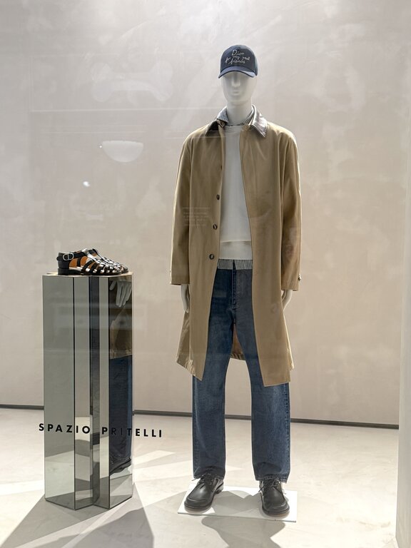 Men's spring outfit in Italy: beige trench coat, dark jeans, light sweater and black leather shoes displayed in an Italian shop window. Photo taken by Lisa Rivera