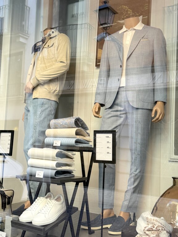 Two men's outfit ideas for Italy: casual jeans and beige jacket alongside a smart casual blue suit with loafers, displayed in an Italian shop window. Photo taken by Lisa Rivera