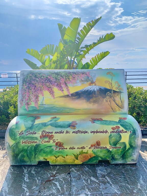 A colourfuly painted bench with a Sicilian saying near the beach at Santa Teresa di Riva, with Mount Etna visible in the distance across the Ionian Sea
